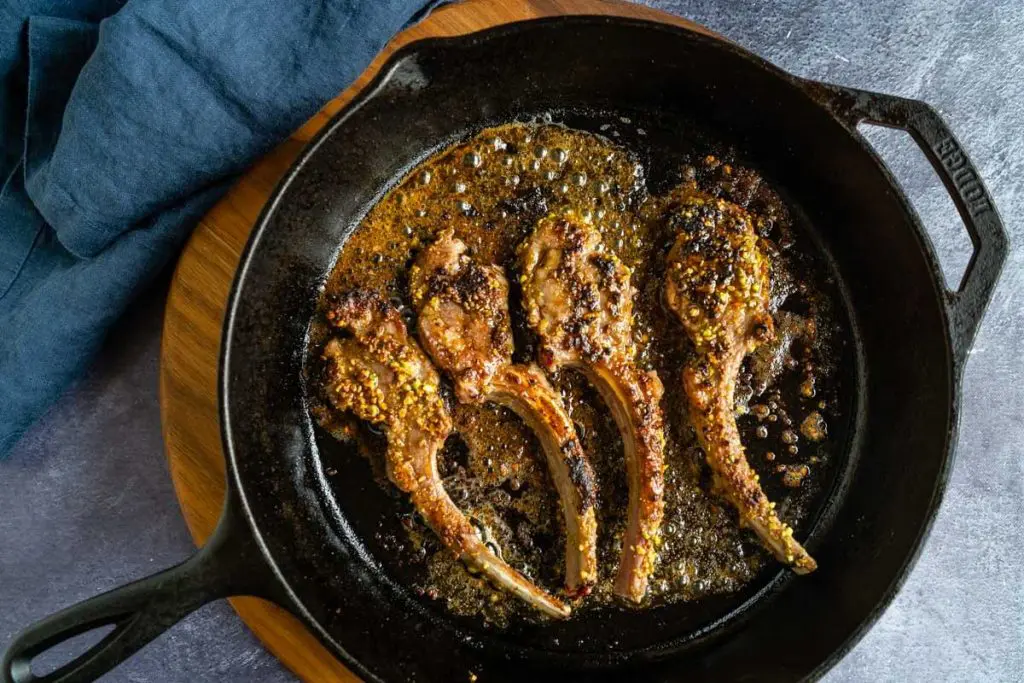 A black cast iron skillet holding sizzling pistachio crusted lamb chops.The skillet sits on a wooden cutting board with a blue napkin in the background over a blue tablecloth.