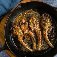 A black cast iron skillet holding sizzling pistachio crusted lamb chops.The skillet sits on a wooden cutting board with a blue napkin in the background over a blue tablecloth.