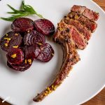 White plate showing a pan-fried pistachio crusted lamb chop next to a serving of sliced red beets sprinkled with pistachios. A sprig of tarragon is on the plate.