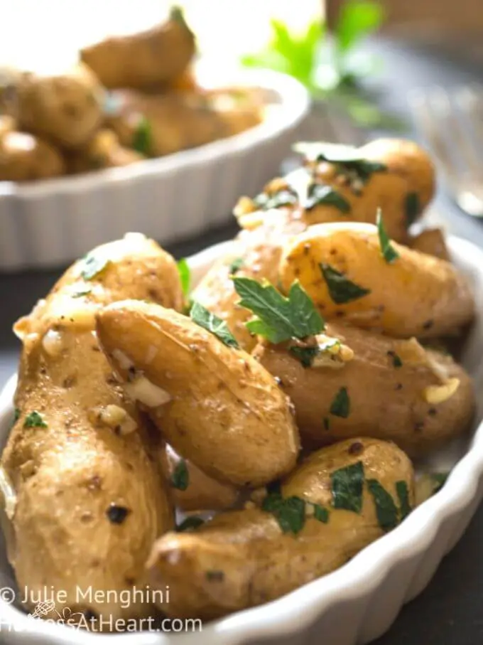 Close up side photo of roasted garlic fingerling potatoes and mined garlic in a white scalloped dish garnished with fresh parsley on a grey slate background. Another bowl of potatoes sit in the background.