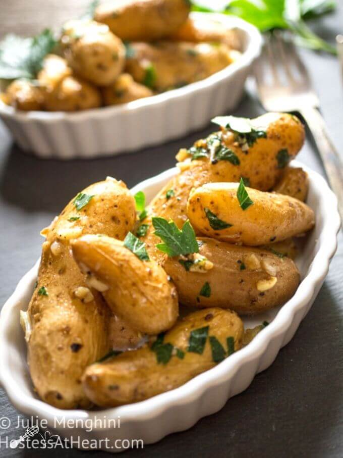 Top angle of a white scalloped dish brimming with roasted stovetop fingerling potatoes and died garlic and garnished with fresh parsley on a slate background and another partial dish of potatoes sit behind it.
