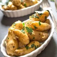 Close up angle view of roasted garlic fingerling potatoes in a white scalloped dish and garnished with fresh parsley on a great slate background. Another bowl of potatoes sit in the background next to a fork.