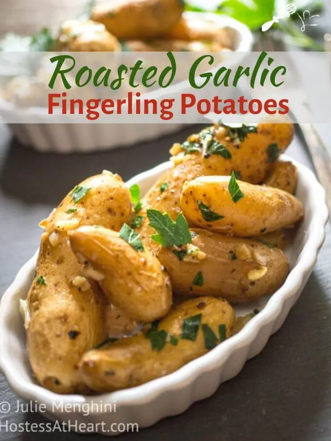 Top angle view of roasted garlic fingerling potatoes in a white dish garnished with fresh parsley sitting on a gray slate background. Another dish of potatoes sits behind it and the recipe title \"Roasted Garlic Fingerling Potatoes\" runs across the top.