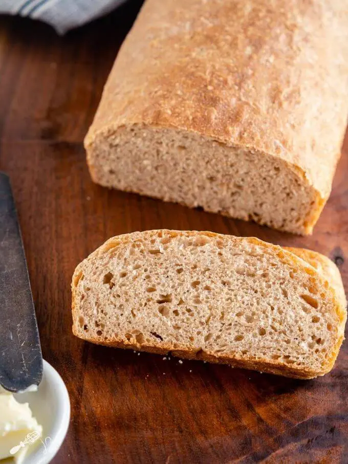 A partial loaf of spelt bread with a slice of bread sitting in front of it on a wooden cutting board. A butter knife and dish of butter sit to the side.