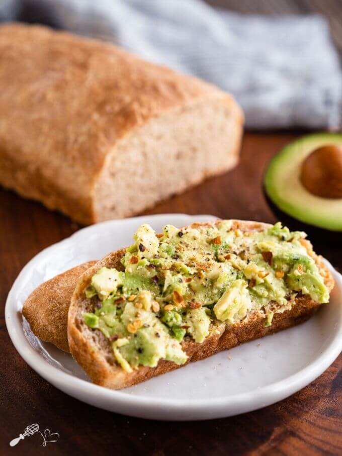 A slice of avocado toast sitting on a white plate over a wooden cutting board. A cut loaf of bread and half of an avocado sit in the background.