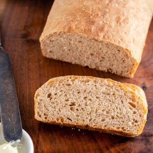 A partial loaf of spelt bread with a slice of bread sitting in front of it on a wooden cutting board. A butter knife and dish of butter sit to the side.