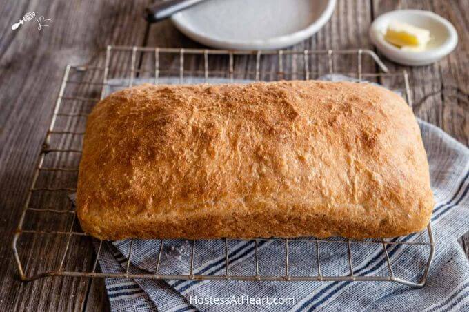 A loaf of browned spelt bread sitting on a cooling rack over a blue striped napkin and wooden background. A pat of butter in a white dish and a white plate sit behind it.