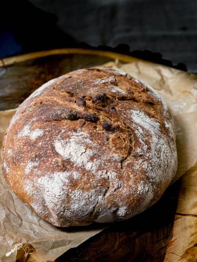 An uncut loaf of spelt bread with a crusty brown exterior dusted with flour. It sits on a piece of natural parchment paper on top of a wooden background.