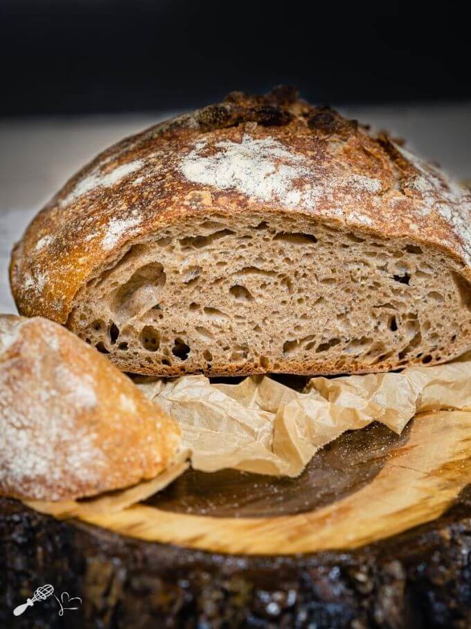 Front shot of a sliced loaf of spelt bread showing a soft crumb and a crusty exterior dusted with flour. The end slice is next to the loaf. The bread sits on parchment paper and a wooden background.