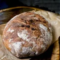An uncut loaf of spelt bread with a crusty brown exterior dusted with flour. It sits on a piece of natural parchment paper on top of a wooden background.