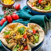 Top angel view of a taco bowl filled with spicy ground chicken and black bean filling topped with shredded cheese, lettuce and tomatoes on a green napkin. A bunch of cherry tomatoes, a bowl of shredded cheese and a partial taco salad sits in the background.