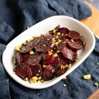 Close-up angle photo of glistening red beet slices in a white bowl sprinkled with chopped pistachio nuts. The dish is sitting on a blue napkin.