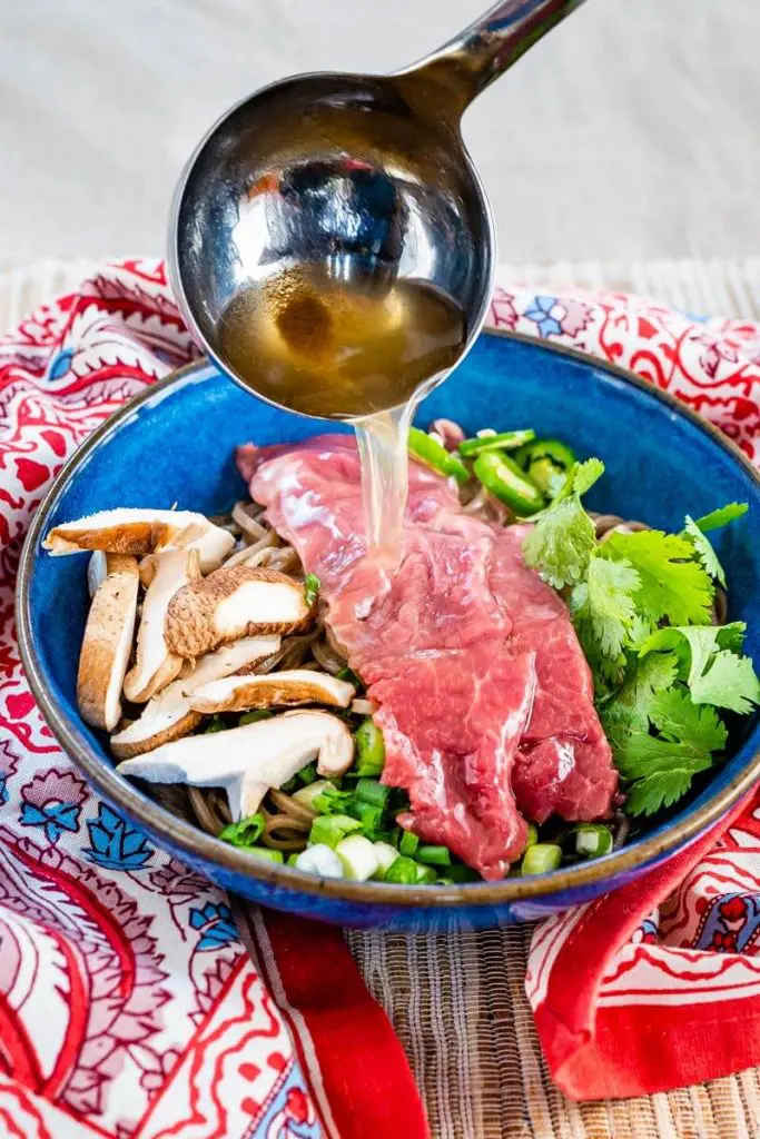 Hot pho broth is being ladled over sliced raw lamb, sliced mushrooms, green onions, Serrano peppers, cilantro, and buckwheat noodles sitting in a blue bowl on a multicolored napkin and grass-cloth placemat.