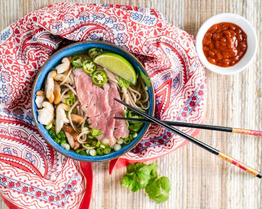 Top down shot of a blue bowl of pho broth loaded with lamb, buckwheat noodles, sliced mushrooms, onions, Serrano peppers, and cilantro. A set of chopsticks are propped on the edge of the bowl that sits on a multicolored napkin next to a bowl of sriracha on a grass-cloth placemat.