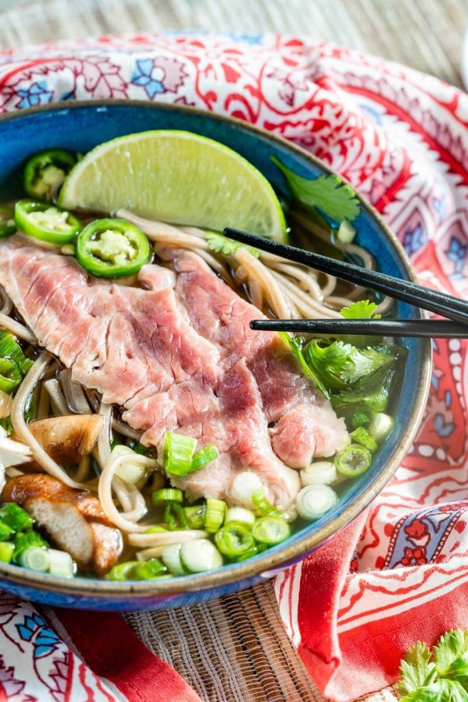 Close-up angle of a bowl of Pho broth over sliced lamb, mushrooms, green onions, cilantro, and Serrano peppers sitting on a multicolored red napkin over a grass-cloth placemat. A wedge of lime is sitting on the back of the bowl waiting to be spritzed over the pho soup.