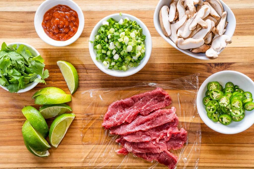 Ingredients used in our homemade pho broth including sliced Serrano peppers, thinly sliced raw lamb, mushrooms, sliced green onion, cilantro, lime wedges, and Sriracha sauce.