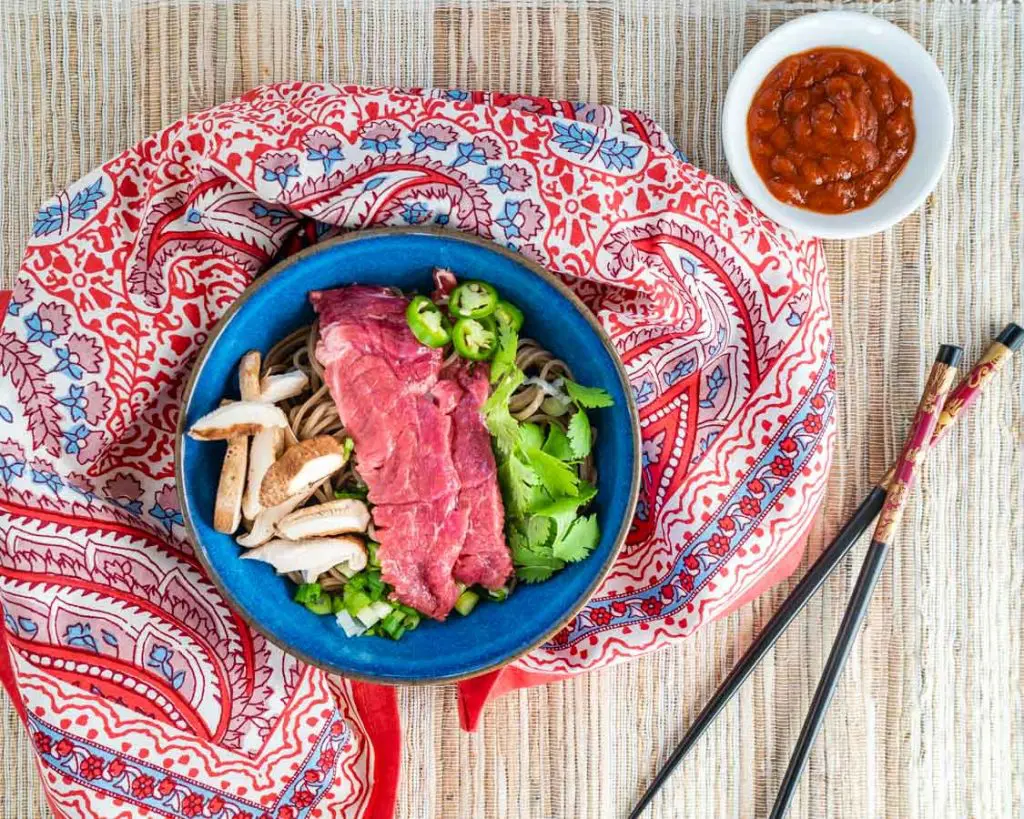 Top down shot of garnishes used in our homemade pho including raw lamb, mushrooms, cilantro, sliced green onions, Serrano peppers, and buckwheat noodles in a blue bowl on a multicolored napkin.. A pair of chopsticks sit to the side and a bowl of sriracha sits in the back.