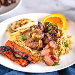 Close-up angle view of chunks of lamb kabob drizzled in an orange glaze sitting on brown rice. Roasted red peppers sit next to the rice on a white plate. A wedge of orange sits in the background.