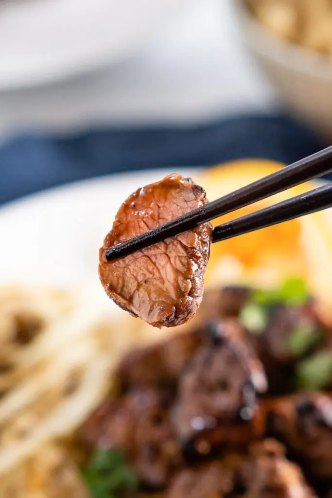A cooked piece of lamb glazed in orange marinade being held with a pair of chopsticks over a blurred serving of lamb kabobs on brown rice.