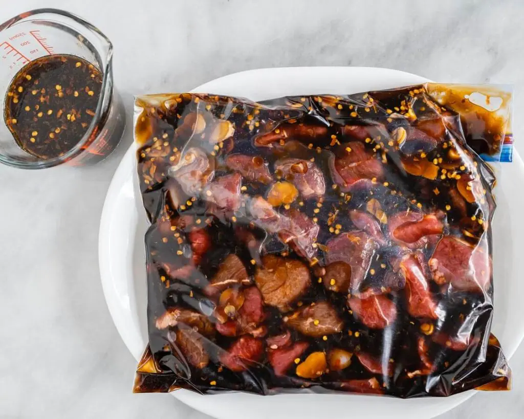 A reclosable plastic bag filled with chunks of lamb marinating in an orange asian sauce. A reserved cup sits next to it.