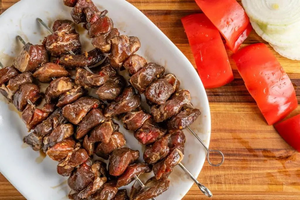Top down view of skewers filled with marinated chunks of lamb on a white plate next to a chopped red pepper and sliced onion. It all sits on a wooden cutting board.