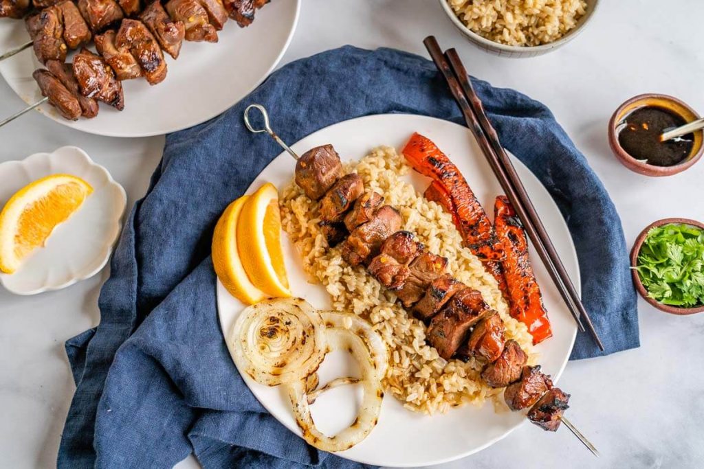 Top down view of a lamb kabob sitting on a bed of brown rice. Roasted onions and red peppers it beside the rice with orange wedges on the back of the white plate which sits on a blue napkin. A set of chopsticks sit on the plate. Small bowls of the orange sauce and cilantro sit beside the plate.