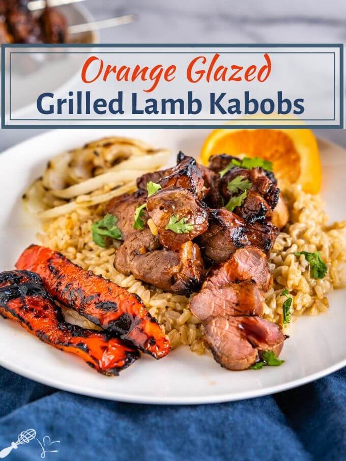 A close-up angle view of chunks of lamb kabob drizzled in an orange glaze sitting on a bed of brown rice. Roasted red peppers and onions sit next to the rice on a white plate. A wedge of orange sits in the background and the title \"Orange Glazed Grilled Lamb Kabobs\" runs across the top.