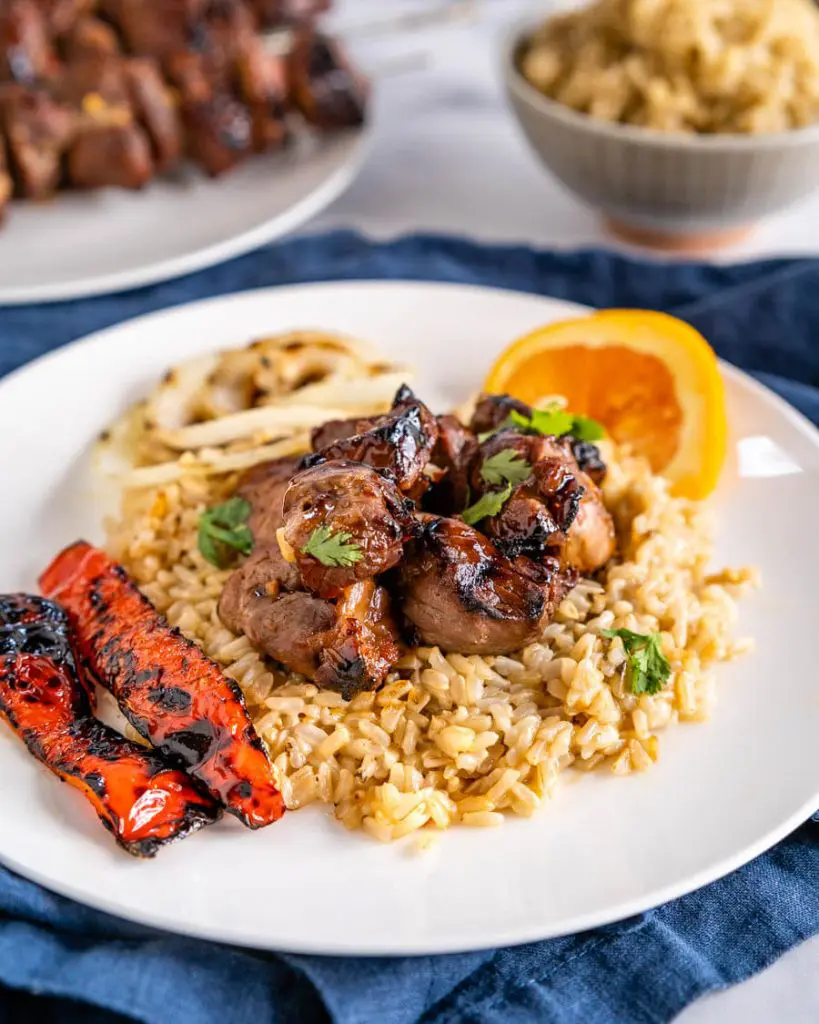 Close-up angle view of chunks of lamb kabob drizzled in an orange glaze sitting on brown rice. Roasted red peppers sit next to the rice on a white plate. A plate of grilled kabobs and a bowl of rice sit in the background.