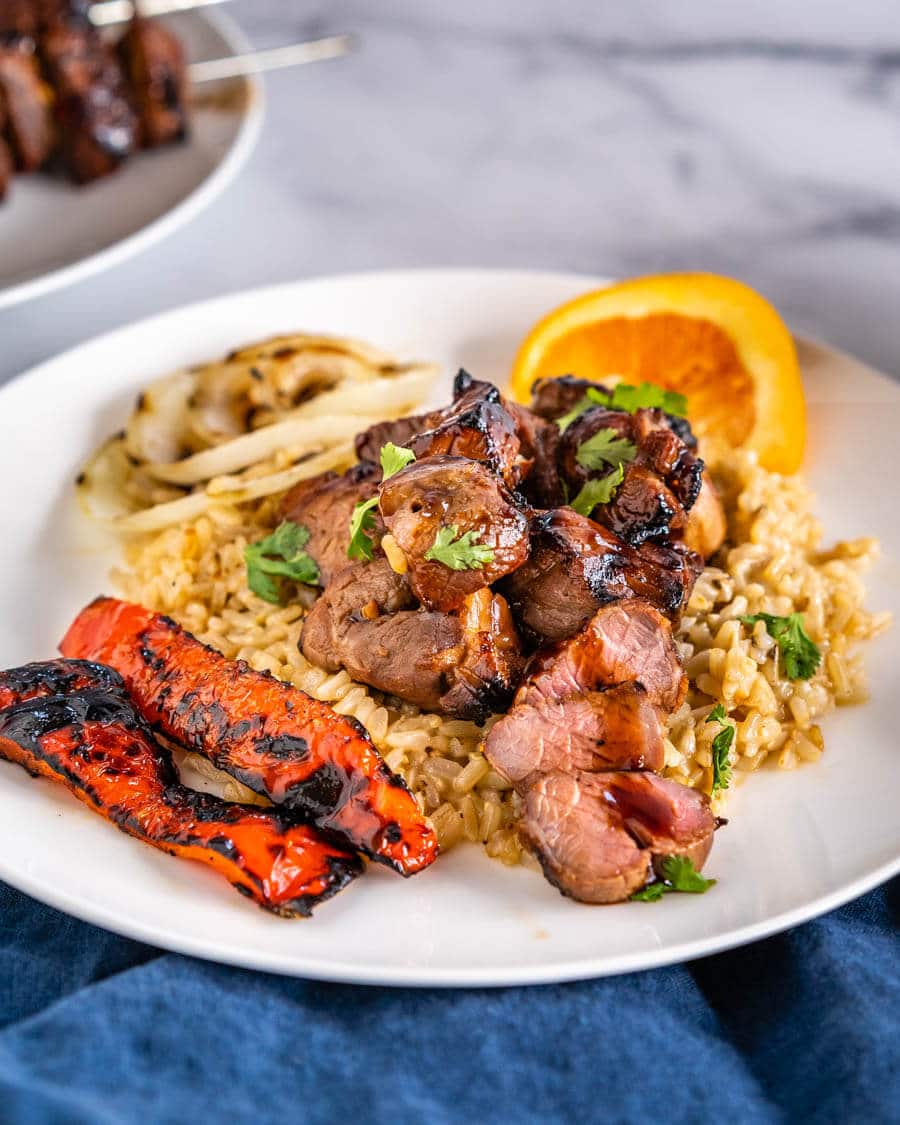 Close-up angle view of chunks of lamb kabob drizzled in an orange glaze sitting on brown rice. Roasted red peppers sit next to the rice on a white plate. A wedge of orange sits in the background.