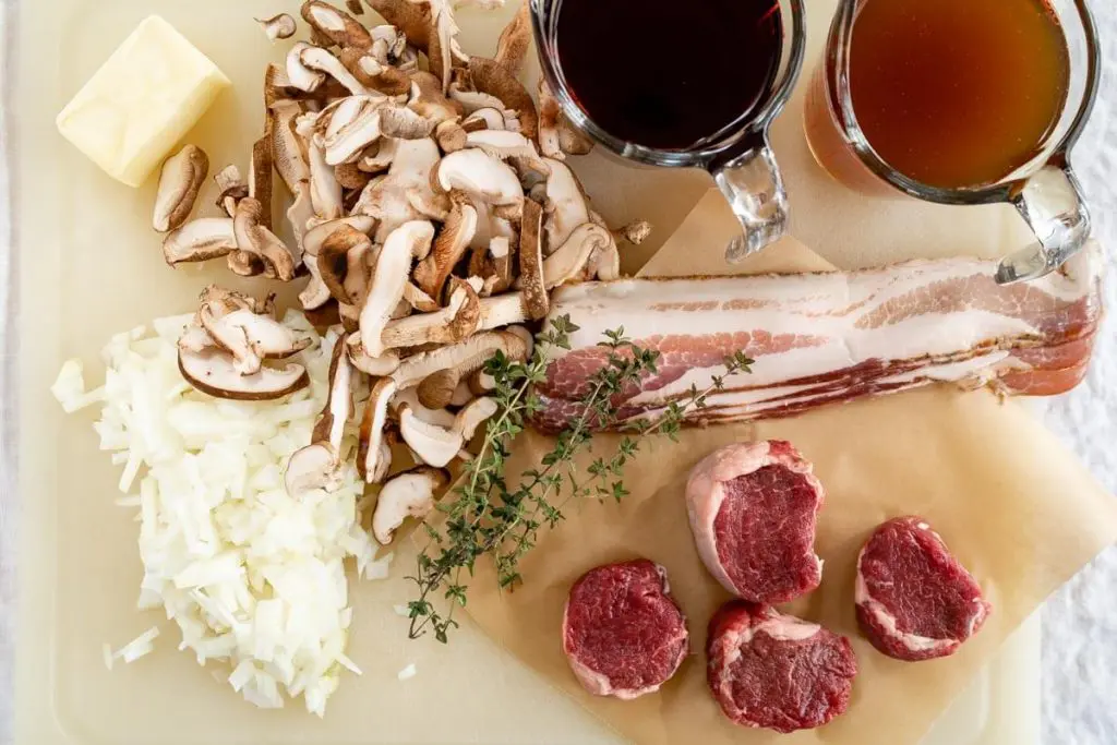 A white cutting board filled with chopped onion, mushrooms, 4 lamb medallions, 4 strips of bacon, 4 tablespoons of butter, fresh thyme, a cup of red wine and a cup of beef broth.
