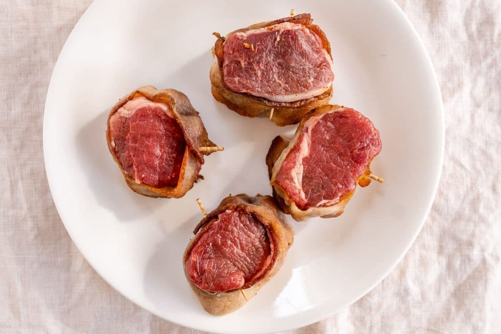 Top down view of 4 lamb medallions wrapped in bacon on a white plate over a white tablecloth.