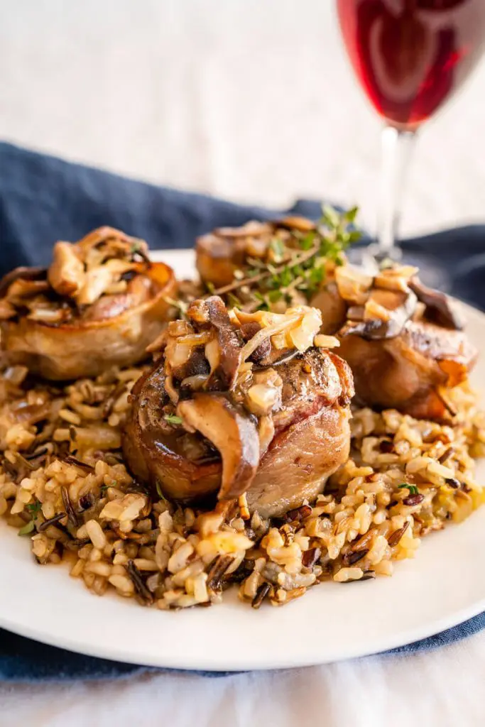 Close-up side view of a white plate with wild rice in the center topped with 4 bacon-wrapped lamb medallions covered in a wine sauce with mushrooms. A sprig of thyme garnishes the dish.