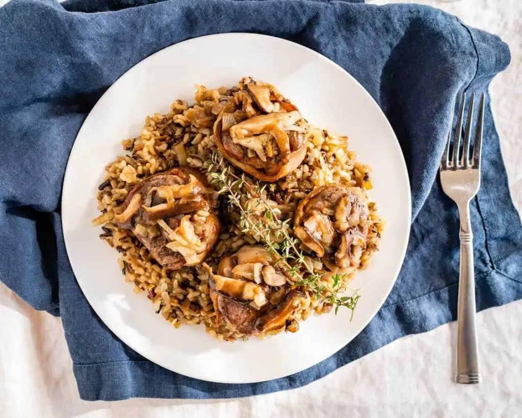 Top down view of 4 bacon-wrapped lamb medallions sitting on a bed of wild rice and topped with a sprig of fresh thyme. on a white plate. A fork sits next to the plate on a blue napkin.