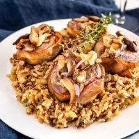 White plate with wild rice in the center topped with 4 bacon-wrapped lamb medallions covered in a wine sauce with mushrooms. A sprig of thyme garnishes the dish.