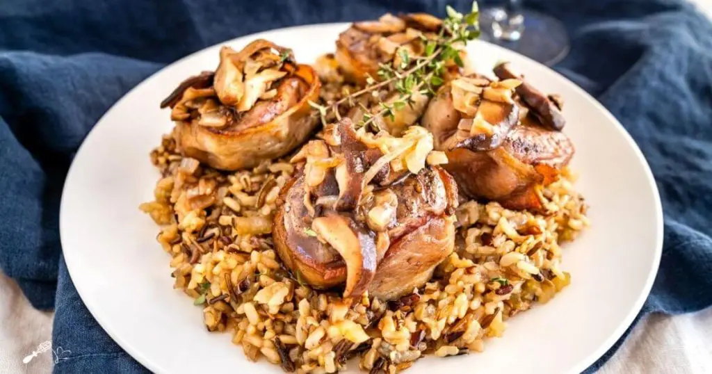 White plate with wild rice in the center topped with 4 bacon-wrapped lamb medallions covered in a wine sauce with mushrooms. A sprig of thyme garnishes the dish.
