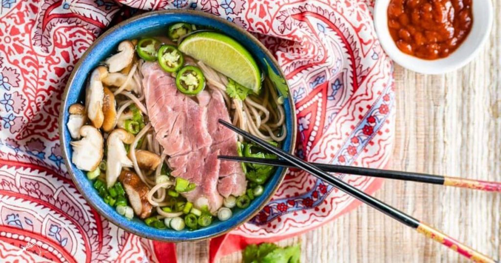 Top down shot of garnishes used in our homemade pho including raw lamb, mushrooms, cilantro, sliced green onions, Serrano peppers, and buckwheat noodles in a blue bowl on a multicolored napkin.. A pair of chopsticks sit to the side and a bowl of sriracha sits in the back.