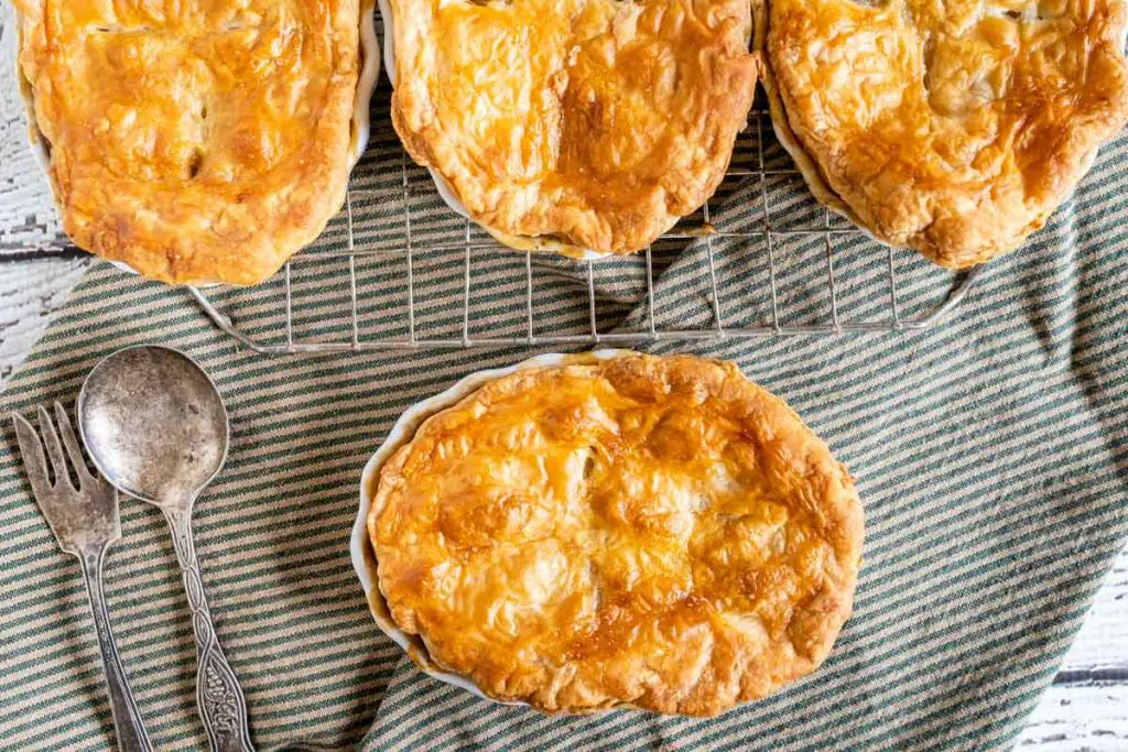 Top down view of ramekin dishes holding Lamb Pot Pies topped with a golden puff pastry crust sitting on a green striped napkin with an antique spoon and fork sitting next to the dish.