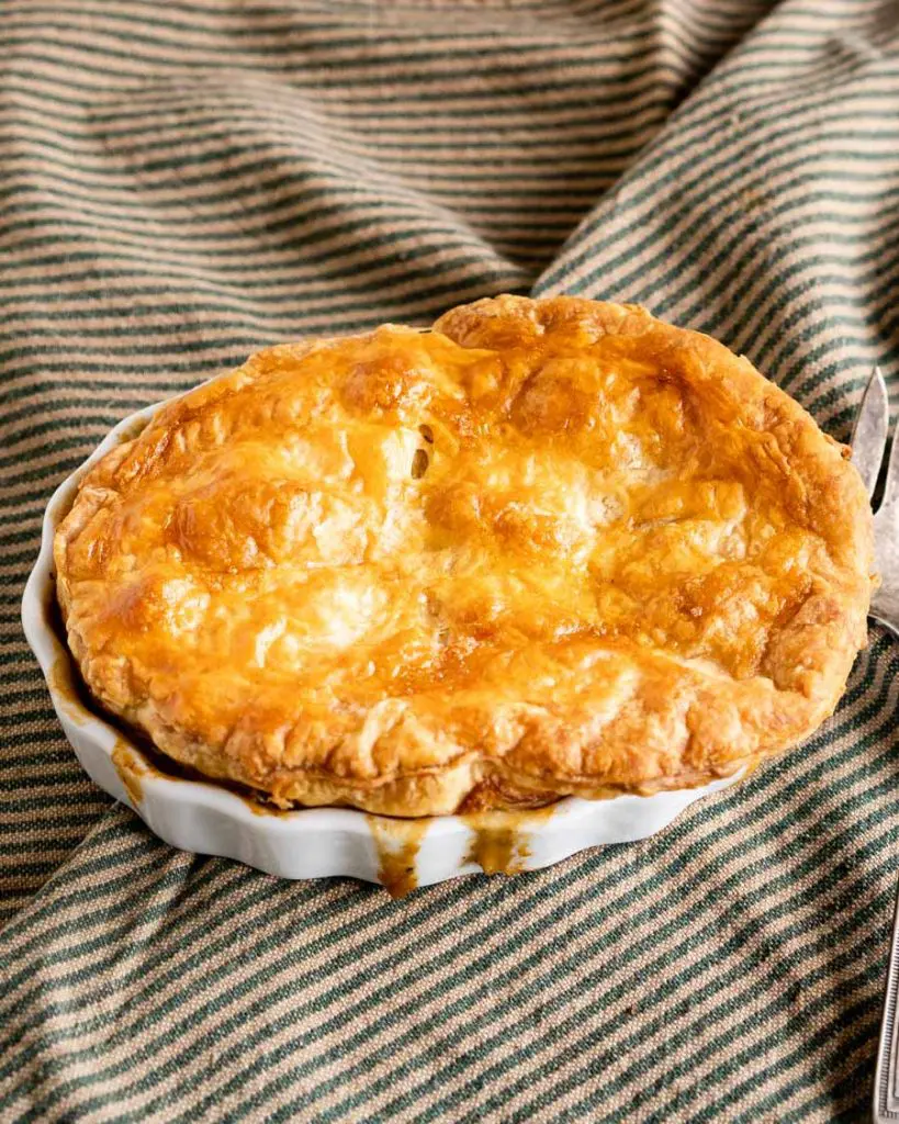 A Lamb pot pie topped with a golden puff pastry crust baked in a white ramekin with the filling dribbling down the sides. The dish sits on a green striped napkin.