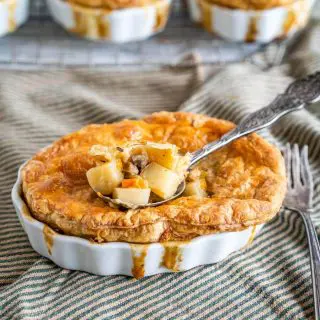 White ramekin filled with a lamb pot pie topped with golden puff pastry and filling spilling down the sides. A spoon of the ingredients is scooped from the center. The dish sits on a green-striped napkin with an antique fork next to the dish.