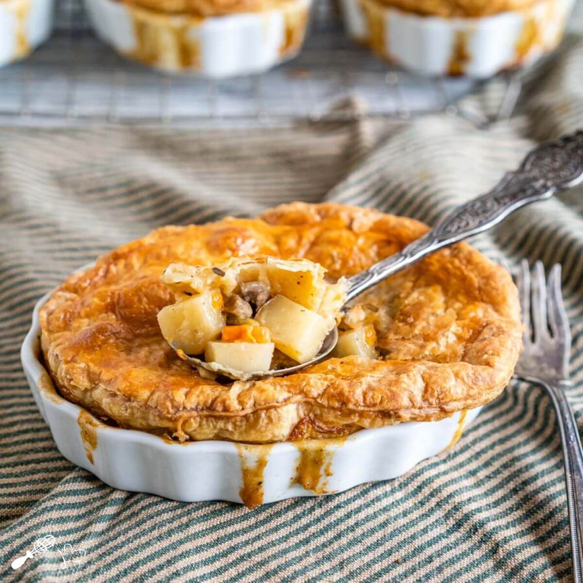 White ramekin filled with a lamb pot pie topped with golden puff pastry and filling spilling down the sides. A spoon of the ingredients is scooped from the center. The dish sits on a green-striped napkin with an antique fork next to the dish.