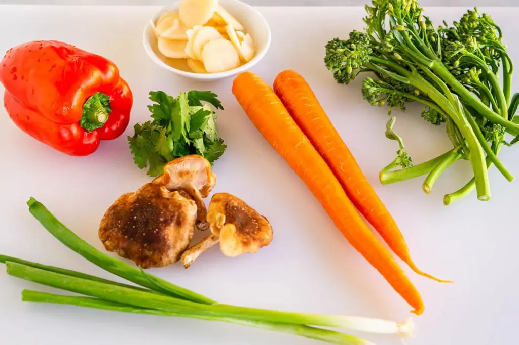 Vegetables in a stir fry including mushrooms, carrots, green onions, broccolini, red pepper and water chestnuts on a white cutting board