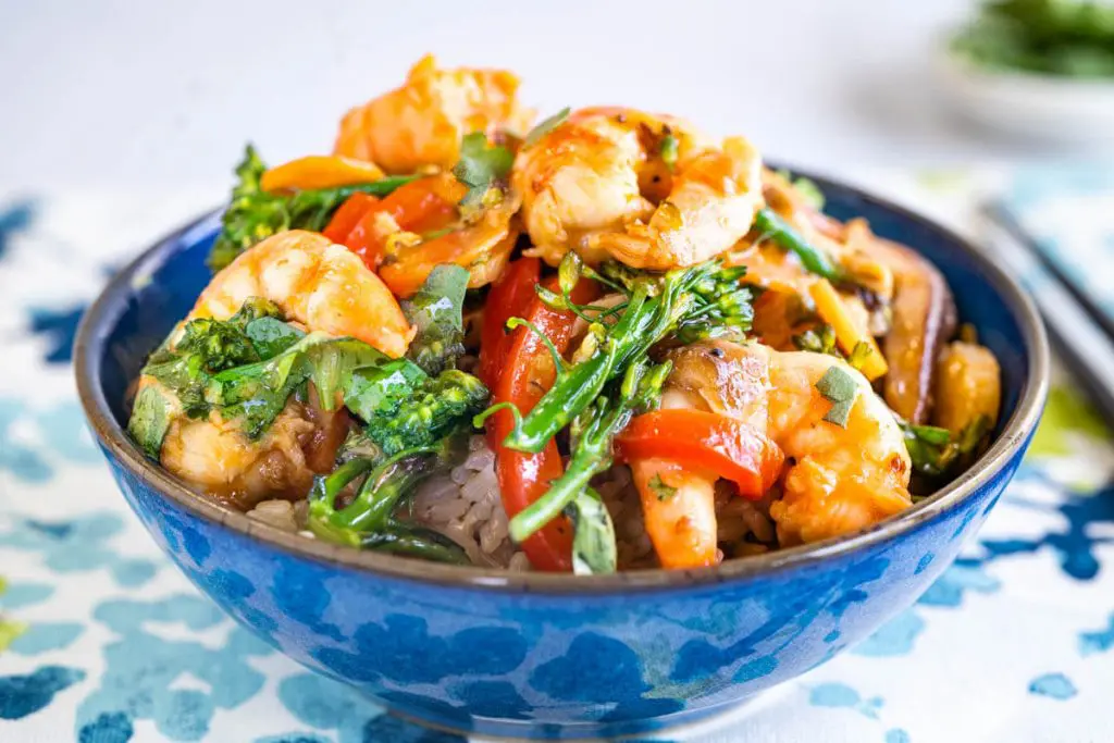 A side view of a blue bowl filled with rice topped with a shrimp stir fry containing broccolini, red peppers, mushrooms, carrots and water chestnuts in a stir fry sauce garnished with fresh cilantro and sitting on a napkin decorated with blue and green designs. A red pan sits in the background.
