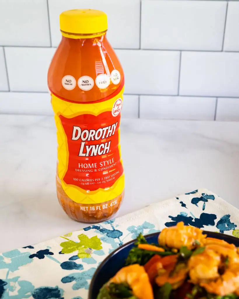 Side view of a bottle of Dorothy Lynch Home Style Dressing and Condiment behind a bowl of shrimp stir fry sitting on a green and blue napkin