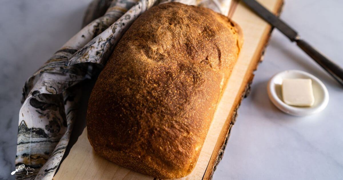 A loaf of sourdough sandwich bread perfectly browned sitting on a cutting board over a piece of marble and a printed napkin.