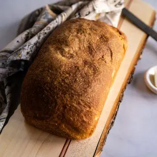 A loaf of sourdough sandwich bread perfectly browned sitting on a cutting board over a piece of marble and a printed napkin. A white dish with butter and an antique knife sit next to it.
