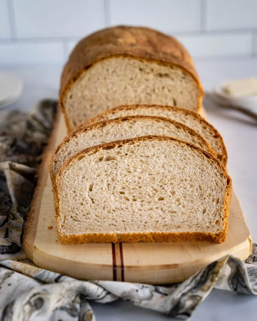 Front angle of a browned loaf of sourdough bread with 3 slices sitting in front of the loaf on a cutting board over a decorative napkin.