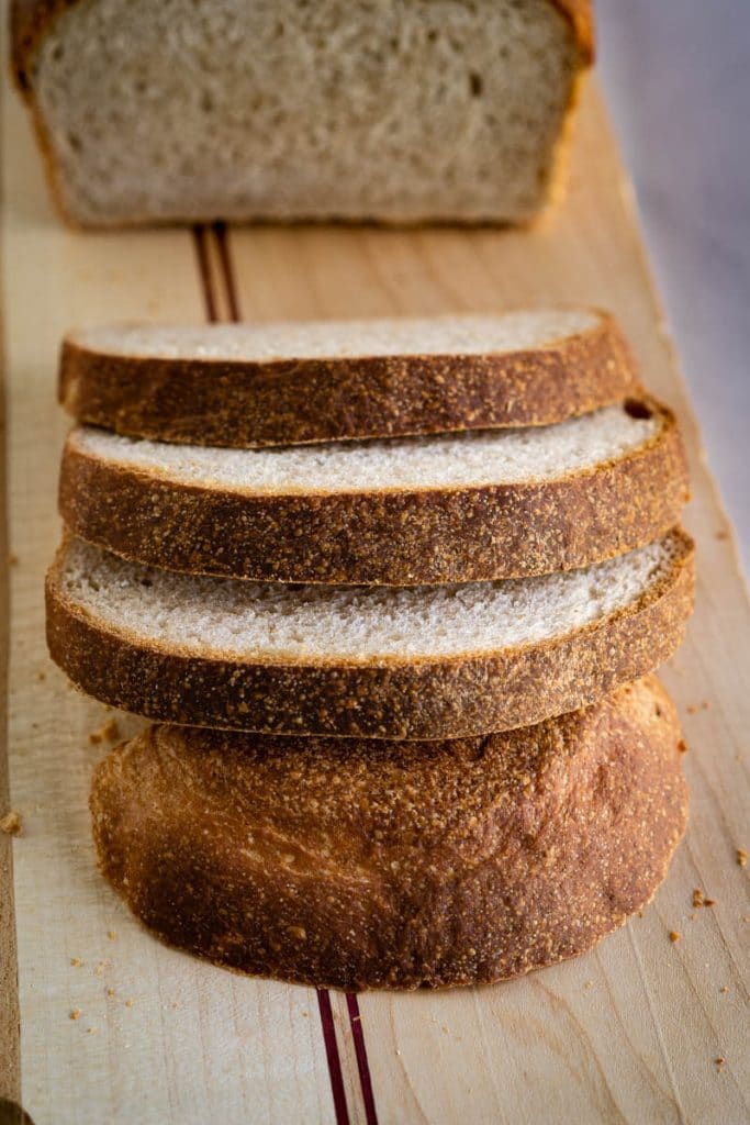 Top angle of 3 slices of sourdough sandwich bread showing golden crust and soft crumb sitting on a cutting board in front of the loaf.