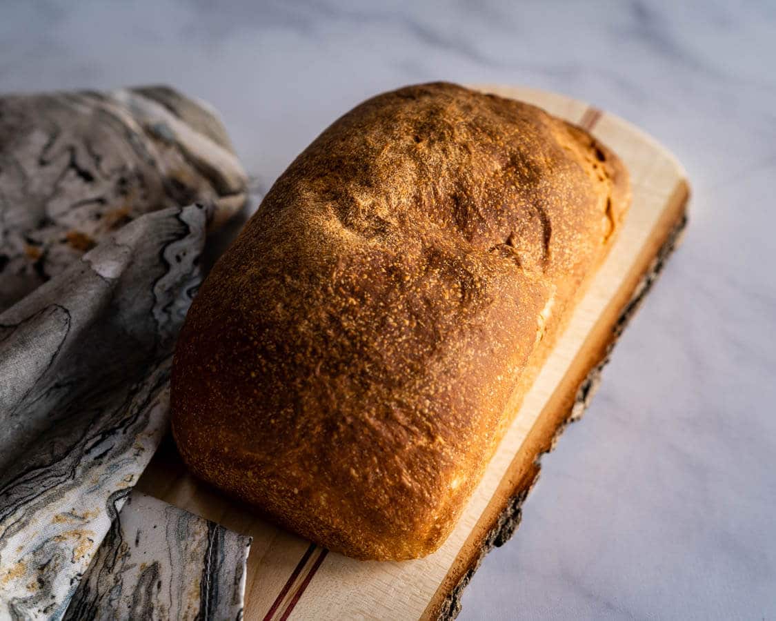 A loaf of sourdough sandwich bread perfectly browned angled on a cutting board over a piece of marble and a printed napkin.