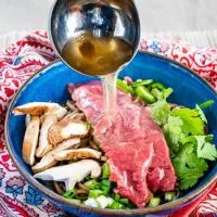 Hot pho broth is being ladled over sliced raw lamb, sliced mushrooms, green onions, Serrano peppers, cilantro, and buckwheat noodles sitting in a blue bowl on a multicolored napkin and grass-cloth placemat.