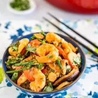 Top angle view of a blue bowl filled with shrimp stir fry containing broccolini, mushrooms, red peppers and water chestnuts sitting on a napkin with blue and green designs. A pair of chop sticks sit in the background,